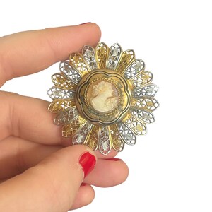 Puede incluir: Broche de camafeo vintage con diseño floral. El centro presenta un retrato de camafeo en un marco dorado. El broche tiene un diseño de filigrana plateada y dorada, creando un efecto dimensional en capas. El broche es redondo.