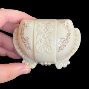 K&ouml;nnte beinhalten: Eine kleine, wei&szlig;e, verzierte Schmuckschatulle mit einem gewellten Rand und braunen Blumenmustern. Die Schachtel hat vier F&uuml;&szlig;e und ist aus Kunststoff.