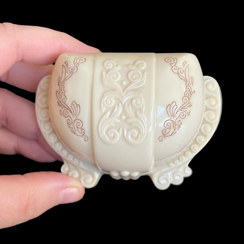 K&ouml;nnte beinhalten: Eine kleine, wei&szlig;e, verzierte Schmuckschatulle mit einem gewellten Rand und braunen Blumenmustern. Die Schachtel hat vier F&uuml;&szlig;e und ist aus Kunststoff.