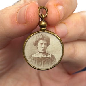 May include: A vintage brass locket with a circular portrait of a woman. The locket has a small loop at the top for a chain. The portrait is in sepia tones, showing the woman's face and upper body.