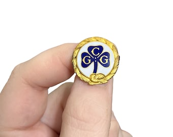 May include: A gold-colored pin with a white clover design and blue lettering that reads "C.G.G."