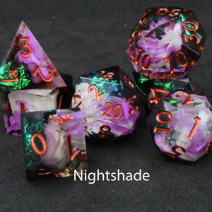 Beautiful black, purple and white resin dice sit on a black felt table top. Light shines down on the dice making the colours swirl into a maelstrom of epic colour. The dice include multicoloured foils that sparkle, shimmer and dance in the light.