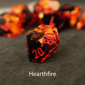 Beautiful black and orange sharp edged dice rest on a black felt tabletop. The dice include a golden foils that shimmer and dance in the sunlight, making it as though flames themselves have been caught inside the dice.