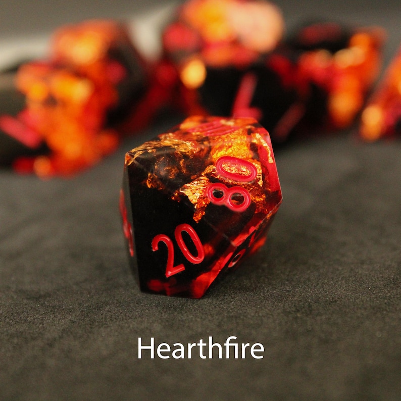 Beautiful black and orange sharp edged dice rest on a black felt tabletop. The dice include a golden foils that shimmer and dance in the sunlight, making it as though flames themselves have been caught inside the dice.
