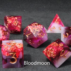 Beautiful red, pink, white and black sharp edge dice rest on a black felt tabletop. The dice include copper and rose gold foils that dance and shine in the sun light.