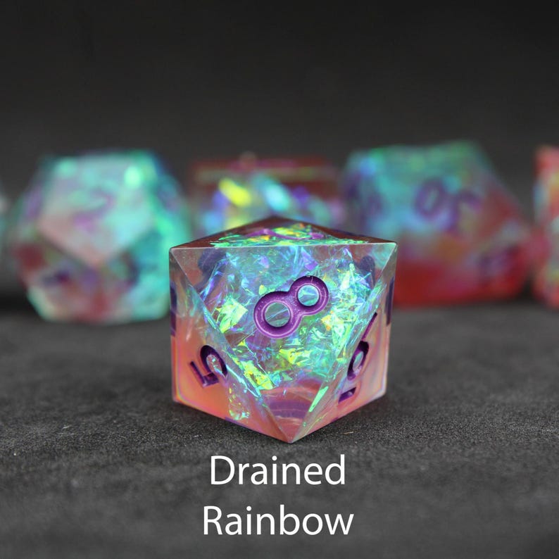 Beautiful red, pink, green and blue sharp edged dice rest on a black felt tabletop. The dice include blue / green foils which shimmer and dance in the sunlight.