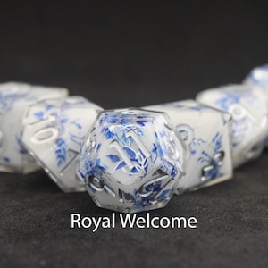 Puede incluir: Juego de dados poliédricos con diseño floral blanco y azul. Los dados tienen números plateados y el texto "Royal Welcome" en la parte inferior. Adecuado para juegos de mesa.
