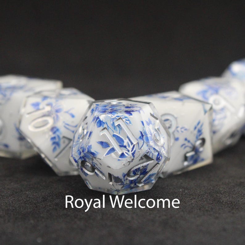 Beautiful white, silver and blue resin dice with gorgeous blue and silver flowers resembling fine china sit on a black felt table top. Light shines down on the dice making the colours pop and sparkle.
