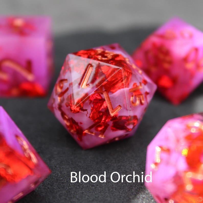 Beautiful pink and purple resin dice inlaid with red holographic flakes rest on a delicate black felt table top. The sun shines down on the red flakes making them shimmer and dance in the light.