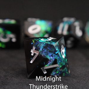 Könnte beinhalten: Nahaufnahme eines schwarzen, vielseitigen Würfels mit schillernden blauen und grünen Einschlüssen. Der Würfel hat weiße Zahlen und den Text "Midnight Thunderstrike" unten. Andere Würfel sind im Hintergrund leicht sichtbar.