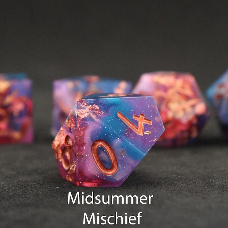 Beautiful blue, pink and purple sharp edged dice rest on a black felt tabletop. The dice include shiny glitter and copper coloured foils that shimmer and dance in the light, creating a wonderful maelstrom of chaotic colour, capturing the true wildness of the feywild.
