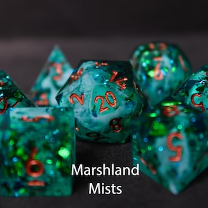 Beautiful blue, white and green edged dice rest on a black felt tabletop. The dice include green and blue foils along with greenish white 'smoke' that swirl within the dice, creating a beautiful maelstrom within.