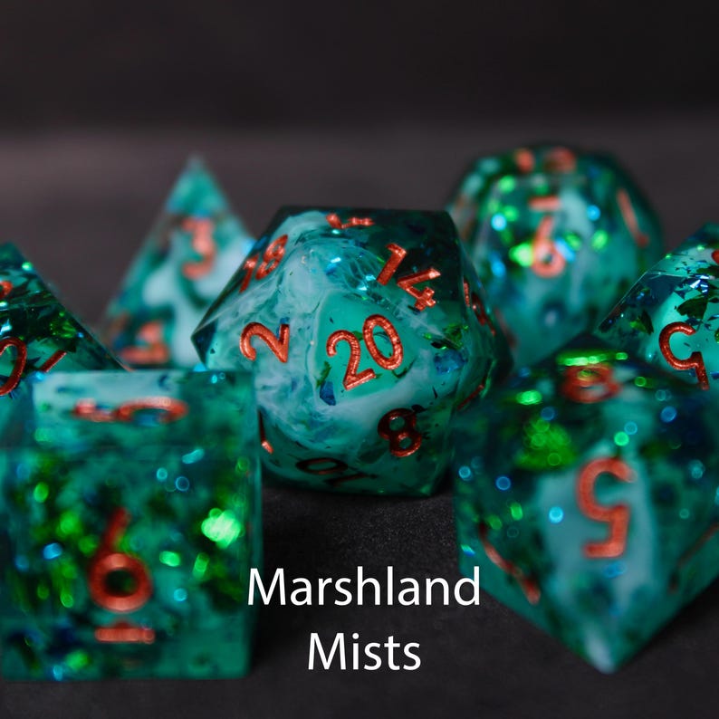 Beautiful blue, white and green edged dice rest on a black felt tabletop. The dice include green and blue foils along with greenish white 'smoke' that swirl within the dice, creating a beautiful maelstrom within.