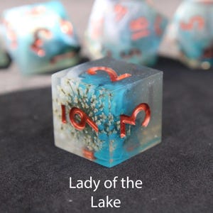 Beautiful blue and white sharp edged dice rest on a black felt tabletop. The dice include a white flower resembling a lily which rests inside the dice, making it appear as if the flower has sunken to the bottom of hauntingly beautiful lake.