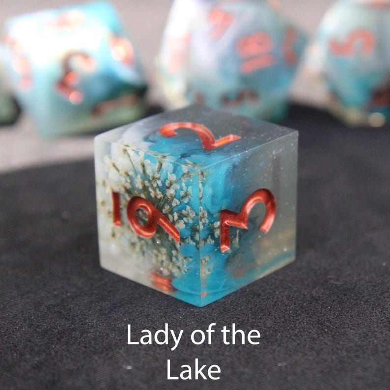 Beautiful blue and white sharp edged dice rest on a black felt tabletop. The dice include a white flower resembling a lily which rests inside the dice, making it appear as if the flower has sunken to the bottom of hauntingly beautiful lake.