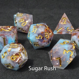Beautiful blue, white and pink resin dice sit on a black felt table top. Light shines down on the dice making the colours swirl into a maelstrom of epic colour. The dice include multicoloured foils that sparkle, shimmer and dance in the light.