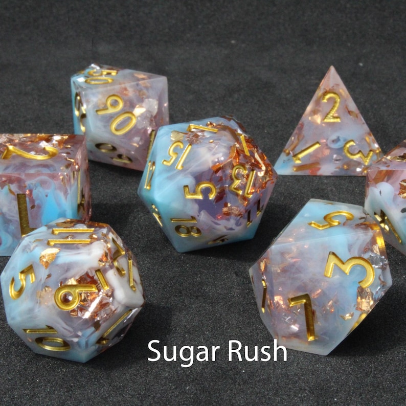 Beautiful blue, white and pink resin dice sit on a black felt table top. Light shines down on the dice making the colours swirl into a maelstrom of epic colour. The dice include multicoloured foils that sparkle, shimmer and dance in the light.