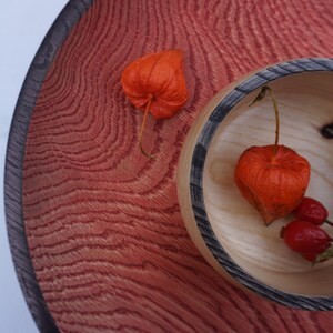 Puede incluir: Un cuenco de madera circular grande con una veta texturizada de color rojo intenso y un borde oscuro. Dentro hay un cuenco de madera más pequeño y claro con borde oscuro, que contiene dos cáscaras de physalis naranjas y dos bayas rojas.