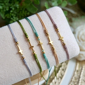 May include: Five adjustable cord bracelets in light gray, olive green, teal, tan, and mauve. Each bracelet features a central gold-toned metal cross charm flanked by two small gold-toned metal beads and knotted cord detailing on both sides.