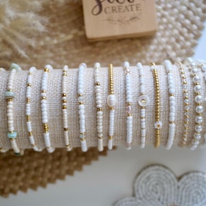 May include: A collection of beaded bracelets in various designs, displayed on a beige fabric roll. The bracelets feature white beads, gold accents, and decorative elements like pearls and small flowers. The bracelets are a mix of delicate and elegant styles.