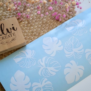 May include: A light blue sheet with white monstera leaf decals. A wooden stamp with the text "Silvi Create" is in the upper left corner. Dried pink flowers and a woven basket are in the background.