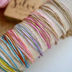 May include: A collection of colorful string bracelets in various shades of pink, gold, blue, orange, and white. The bracelets are displayed on a beige fabric roll, with a blurred background. The bracelets are made of thin, woven threads.