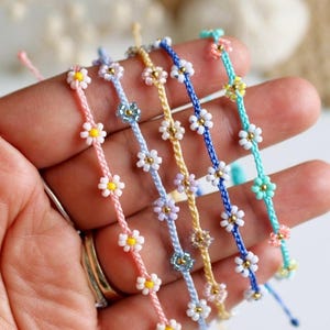 May include: A collection of daisy chain bracelets in various colors, including pink, blue, yellow, and turquoise. Each bracelet features small white and yellow daisy beads strung along a cord. The bracelets are delicate and suitable for everyday wear.