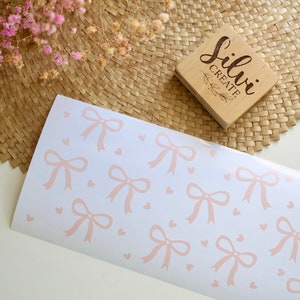 May include: White paper with a repeating pattern of light pink bows and hearts. A wooden stamp with the text "Silvi Create" is visible. Dried pink flowers and a woven mat are in the background.