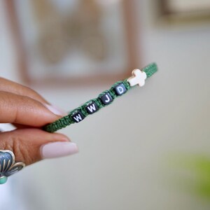 May include: A green woven cord bracelet featuring four black square beads with the white letters W, W, J, and D printed on them, followed by a small white cross-shaped bead. The bracelet is constructed with a simple knot pattern.