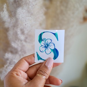 May include: A white sticker featuring the letter "S" with a blue and green marbled design. A blue hibiscus flower is integrated into the letter's design. The sticker is held by a hand with a gold ring.