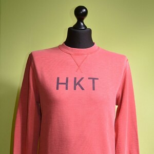 Puede incluir: Sudadera de color coral con las letras "HKT" impresas en gris oscuro en el pecho. La parte superior de manga larga tiene cuello redondo con un detalle cosido y se muestra sobre una cabeza y un soporte de maniquí negros.