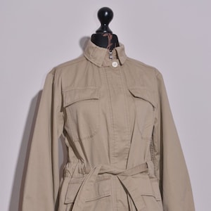 Pode incluir: Uma jaqueta utilitária bege com cinto, dois bolsos grandes com aba e fecho de correr.