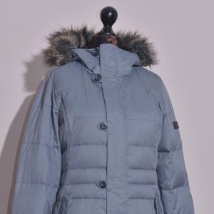 Puede incluir: Chaqueta acolchada azul claro con capucha ribeteada de piel. La chaqueta tiene cierre de botones, dos bolsillos delanteros y un diseño acolchado. La chaqueta se exhibe en un maniquí negro.