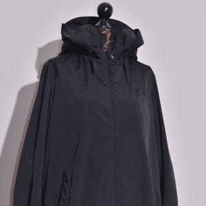 Può includere: Un impermeabile nero con cappuccio, chiusura a zip e tasche. Il cappotto lungo è realizzato in un materiale liscio e resistente all'acqua. Il cappuccio è attaccato e ha un cordoncino. Il cappotto è esposto su un manichino.
