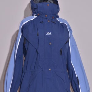 May include: A navy blue hooded jacket with light blue sleeves and white accents. The jacket features a drawstring waist, multiple pockets, and the "HH Helly Hansen" logo. It is displayed on a black mannequin.