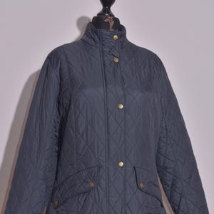 May include: A dark navy quilted jacket with a stand-up collar and gold-colored buttons. The jacket has two front pockets and a diamond pattern throughout. The jacket is displayed on a black mannequin.