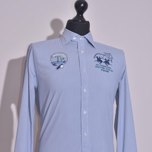 Op de afbeelding: Lichtblauw overhemd met verticale strepen en knopen. Voorzien van geborduurde patches op de borst met tekst en paardensportafbeeldingen. Lange mouwen met manchetten met knopen. Het overhemd wordt op een mannequin getoond.