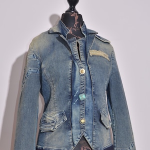 Peut inclure: Veste en jean bleu clair avec une coupe ajustée, col, fermeture boutonnée et deux poches avant. La veste présente des coutures décoratives et des boutons dorés. La veste est présentée sur un mannequin.
