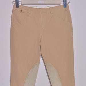 Puede incluir: Pantalones de montar color beige con parches de ante en las rodillas y detalles de botones en los tobillos. Los pantalones cuelgan de una percha sobre un fondo blanco. Diseño clásico y a medida.