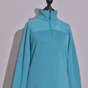 May include: A turquoise fleece pullover with a high collar and a quarter-zip closure. The long-sleeved top features a small logo on the chest and thumb holes at the cuffs. The garment is displayed on a mannequin.