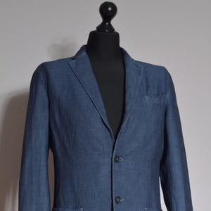 Puede incluir: Un blazer azul con dos bolsillos delanteros, un bolsillo en el pecho y dos botones. El blazer se muestra sobre una cabeza y torso de maniqu&iacute; negros, sobre un fondo neutro. La tela tiene una textura sutil.