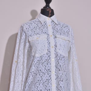 May include: White lace button-down shirt with long sleeves and two chest pockets. The shirt is displayed on a black mannequin. The lace pattern features a floral design. The shirt has a classic collar and buttoned cuffs.