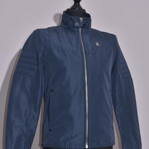 May include: A navy blue jacket with a high collar and a full-length silver zipper. The jacket has quilted detailing on the shoulders and a small logo on the chest. It is displayed on a black mannequin.