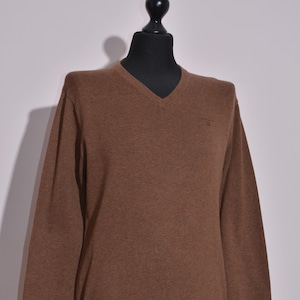 Puede incluir: Un suéter marrón con cuello en V exhibido en un maniquí. El suéter de manga larga tiene dobladillo y puños acanalados. El logotipo de la marca es visible en el pecho. El suéter está hecho de un material de punto suave.