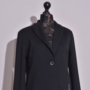 Puede incluir: Una chaqueta negra con un solo botón de cierre y un detalle de puntada decorativa en el lado derecho. La chaqueta tiene un cuello de muesca y mangas largas.