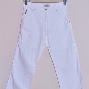 Może przedstawiać: Białe spodnie stolarskie z wieloma kieszeniami i prostą nogawką. Spodnie wykonane są z wytrzymałego materiału i mają klasyczny pięciokieszeniowy design, z małą kieszenią po prawej stronie. Spodnie wiszą na wieszaku.