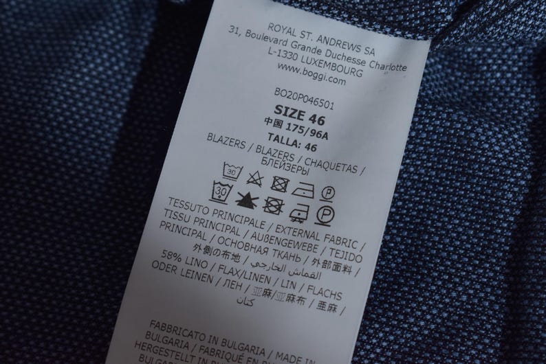 Puede incluir: Primer plano de una etiqueta de ropa blanca con texto negro, incluyendo la talla 46. La etiqueta est&aacute; adherida a una tela azul marino texturizada. La etiqueta tambi&eacute;n incluye instrucciones de lavado y la composici&oacute;n de la tela.