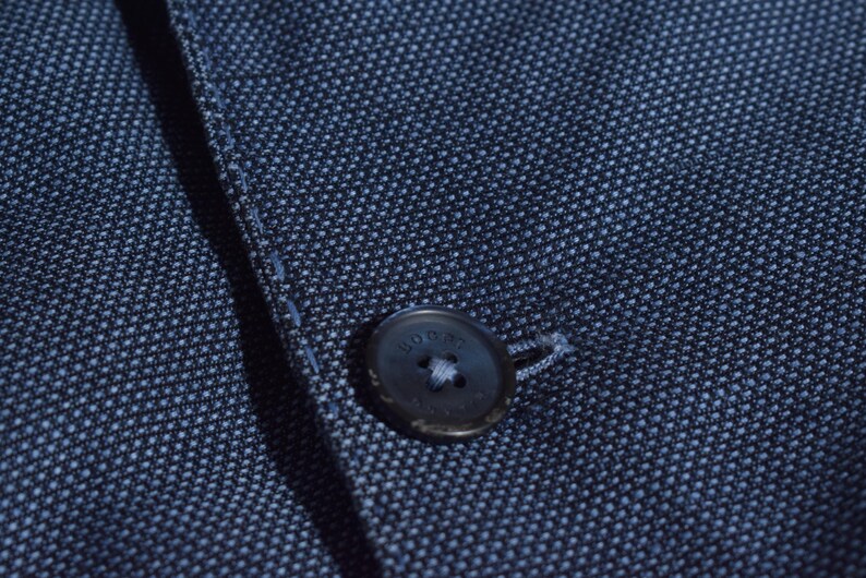 Puede incluir: Primer plano de una blazer azul marino con tejido texturizado. Se ve un bot&oacute;n oscuro con la palabra "HOGAN", sujeto con hilo blanco. La tela tiene un sutil estampado de puntos.