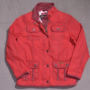 Könnte beinhalten: Rote Jacke mit vier Vordertaschen, Reißverschluss und Knopfleiste. Die Jacke hat einen Cordkragen und ein floral gemustertes Futter. Auf der Vordertasche befindet sich ein kleines Logo.