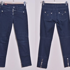 Puede incluir: Vaqueros azul marino oscuro con cremallera y tachuelas plateadas. Los vaqueros presentan un diseño ajustado con paneles en las rodillas y detalles de botones en los tobillos. La vista trasera muestra dos bolsillos con cierres de botón.